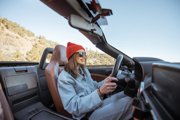 Happy woman dressed casually in jacket and red hat driving convertible car on the beautiful...