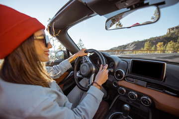 Happy woman dressed casually in jacket and red hat driving convertible car on the beautiful...