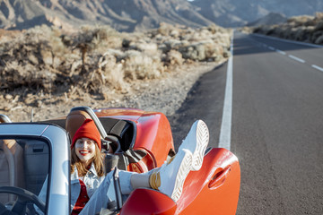 Young joyful woman traveling by convertible car on the desert valley, pulling legs out of the car window on the roadside. Lifestyle in travel concept