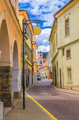 Narrow street in Kutna Hora historical Town Centre with cobblestone road, old style colorful buildings and street lights lamp, blue cloudy sky background, Central Bohemian Region, Czech Republic