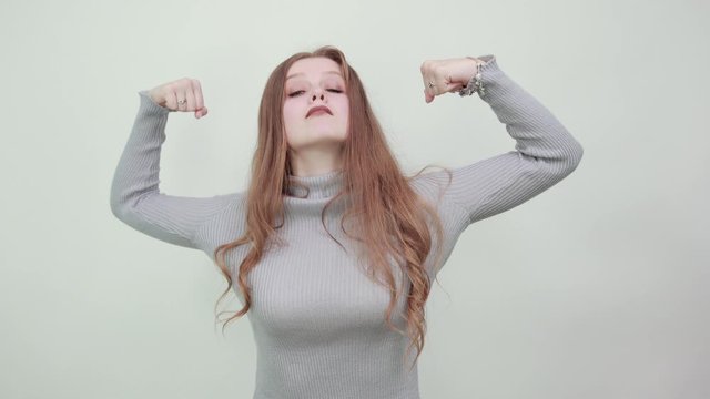 A young beautiful red haired woman in gray sweater shows her muscles on arms demonstrating strength confidence power