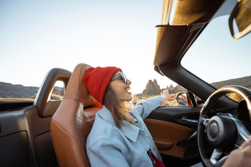 Happy woman in red hat driving convertible car while traveling on the desert road with beautiful...