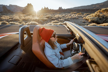 Happy woman in red hat driving convertible car while traveling on the desert road with beautiful...