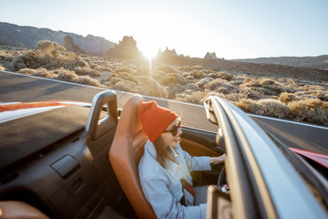 Happy woman in red hat driving convertible car while traveling on the desert road. Image focused on...