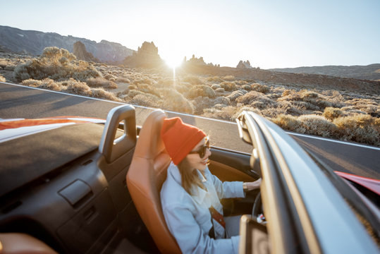 Happy woman in red hat driving convertible car while traveling on the desert road. Image focused on the rocks on the background