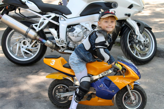 A little kid child girl biker in motorcycle protection short jean skirt on a yellow with a blue children's electric motorcycle minimoto stands next to an adult large motorcycle child 