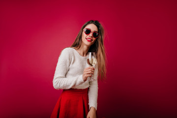Inspired girl with wineglass in hand looking to camera. Portrait of interested brown-haired woman in heart sunglasses isolated on claret background.