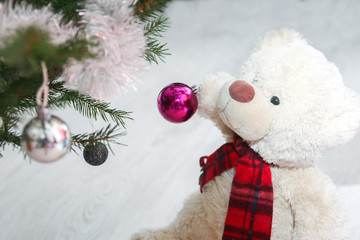 Fototapeta na wymiar A cute teddy bear with scarf is giving the christmas decoration on the christmas tree. He holds the ball in its paws. 