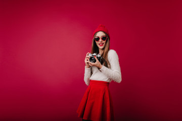 Obraz premium Indoor portrait of interested girl in sunglasses and red hat isolated on claret background. Glamorous female photographer in skirt standing in studio with camera.