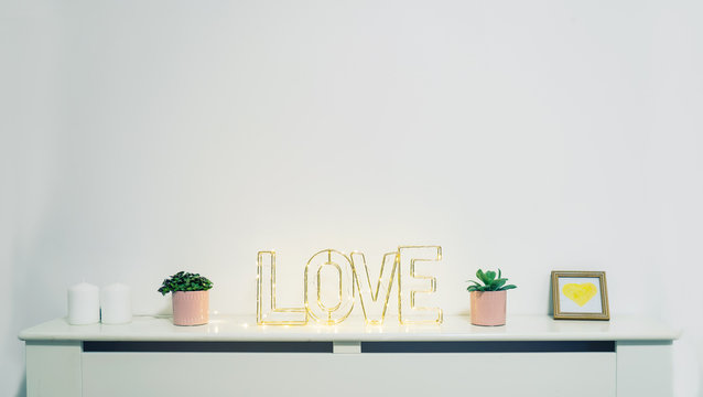 Home interior decoration with wire word Love in lighting garland and home potted flowers and candles near white wall. Valentine's day concept. Romantic mood in simle minimalism style. Copy space
