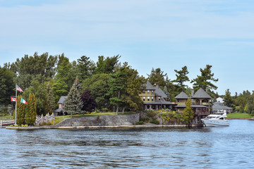 Obraz premium Building in a grassy area on the coast of a lake during daytime, and surrounded by trees. Yacht parked in the lakeside harbor. Real state concept. Thousands Islands. Ontario, Canada