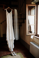 Fototapeta na wymiar Hanging classy wedding dress and wedding veil in the room near the opened window