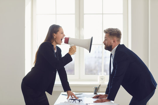 Angry businesswoman shouting into a megaphone at a worker man standing at a table in the office.