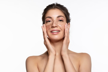 Beautiful woman with perfect skin applying exfoliating coffee scrub on nose and cheeks studio...