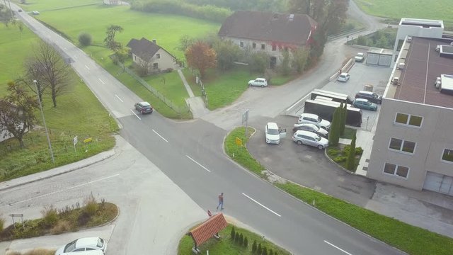 An aerial view of the traffic moving at a square road in a small town having lush greenery and vehicles parked at the building around.