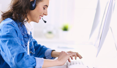 Happy charming young woman sitting and working with laptop using headset in office