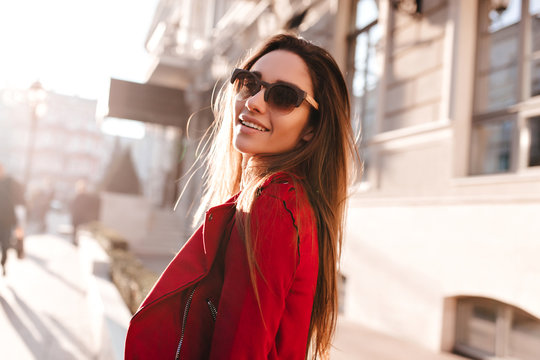 Portrait of cute long-haired girl in sunglasses looking over shoulder on blur city background. Outdoor photo of joyful caucasian woman in casual red outfit.