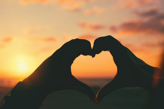 Girls hands in heart shape with sun light and colorful sky on the beach as a background.