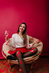 Blissful girl sitting in cozy chair under heart confetti and smiling. Lady with long hairstyle wears white sweater drinking champagne in holiday.