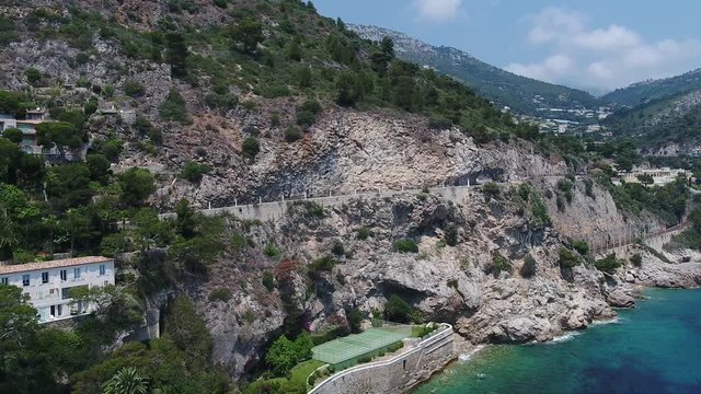 Car Driving On The Highway In Antibes, France Along The Lush Rocky Mountain Near The Blue Sea Water On A Sunny Day - Aerial Shot