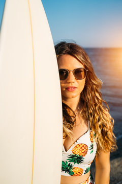 Close up of an attractive young woman wearing sunglasses and wearing Hawaiian clothes holding a surfboard on the beach - Vertical image