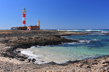 View over beach and rocky ground on isolated red and white striped lighthouse contrasting against blue sky in north of Fuerteventura