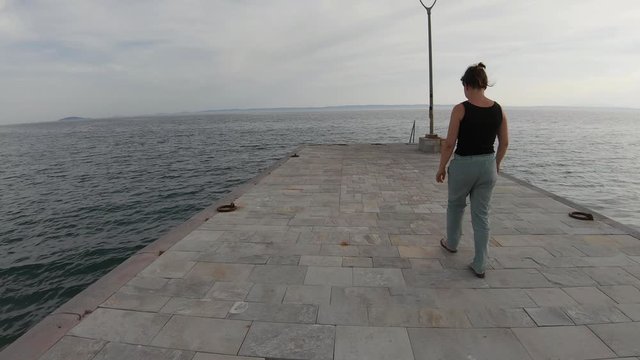 Brunette girl walking on a sea dock in Greece. Back following view with tranquillity horizon in fromt of her.