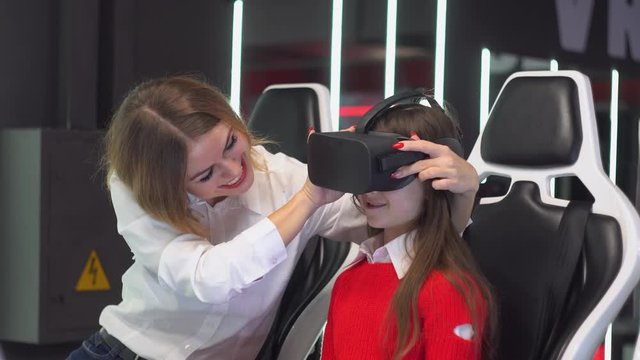A woman puts glasses on her daughter. Woman and little girl are getting ready to start playing virtual reality simulator