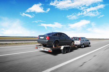A crossover with trailer towing a car on the countryside road in motion against sky with clouds