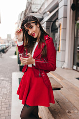 Fototapeta premium Lovable dark-haired girl in red skirt enjoying outdoor photoshoot. Excited young female model in stylish jacket holding cup of tea on the street.