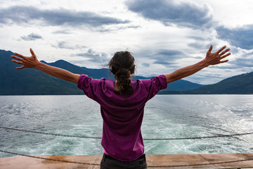 Close up from behind of a man on the deck of a passenger ferryboat. Arms wide open in enthusiastic appreciation of the landscape. Freedom concept.