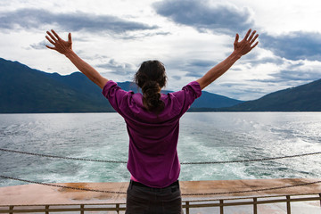 Close up from behind of a man on the deck of a passenger ferryboat. Arms raised in enthusiastic...