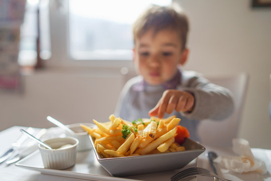 Portrait of small little cute caucasian boy kid eating french fries potato chips at the table in the restaurant or at home three or four years old focus on potatoes