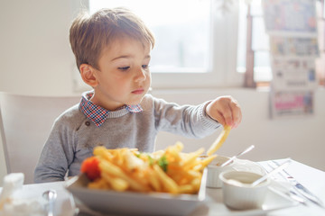 Portrait of small little cute caucasian boy kid eating french fries potato chips at the table in...