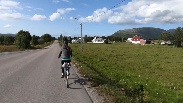 A young woman is biking on an old retro bike on the side of a countryside road in the north of Norway within the arctic circle. Green grass fields on the side with small houses.