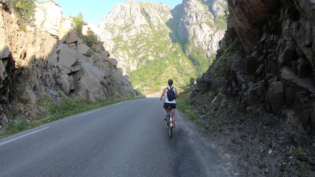 A cute, young woman is biking through a passage on an old retro bike with a small backpack on the side of the road with large and steep mountains ahead of her in Lofoten, Norway.