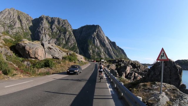 A cute, young woman is biking uphill on an old retro bike with a small backpack on the side of the road with the epic scenery of north Norway's mountains around her. Sunny day near Henningsvaer.