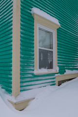 The exterior corner of a green building with a small window.  The trim on the building and around the window is beige in color. There's a snow drift on the ground and snow on the window frame. 
