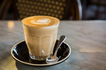 Coffee latte in glass cup with love heart shape latte art in milk froth, on a marble table and retro chair background in a coffee shop