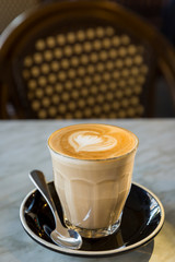 Coffee latte in glass cup with love heart shape latte art in milk froth, on a marble table and retro chair background in a coffee shop, vertical view