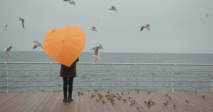 Woman in parka with orange umbrella in heart shape enjoying sea view with seagulls in storm. Overcast weather.