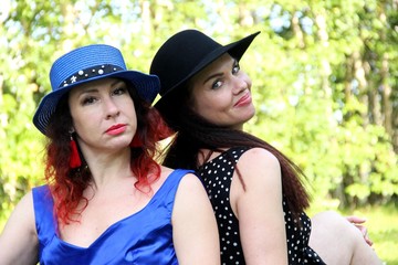 Two female friends sit back to back on a green lawn on a Sunny day. a red-haired woman in a blue hat and dress, a brunette in a black hat and dress, close-up