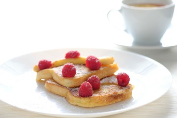 Two sweet toasts with raspberries sprinkled with sugar served on a white plate with tea in the background.