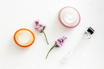 Fototapeta na wymiar Cosmetologist tools. Dermaroller, creams near leaves and flowers on white background top-down flat lay