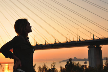 Fototapeta premium the silhouette of a young beautiful girl with her hands on her waist looks at a large cable-stayed bridge on the background of the sunset, a moment of enjoying life in a big city, matte mood toning