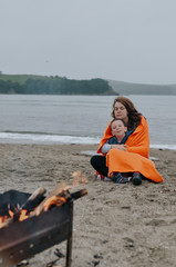 mother and son covered with an orange fleece blanket sit on the sandy beach and warm themselves by the fire on a foggy cold summer day, strong tender relations between generations and family