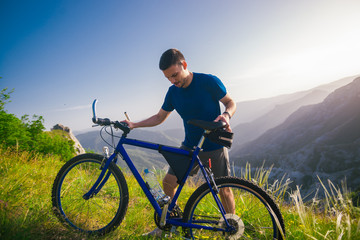 Obraz premium Perspective of a fit mountain biker pushing his bike uphill with amazing view on a forest, river and mountains in the background. Amazing green nature at sunset.