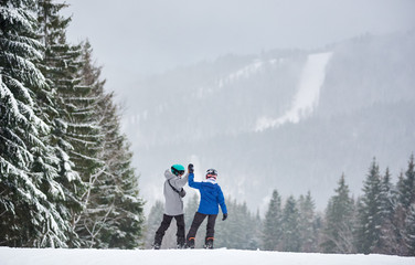 Couple of snowboarders with hands up preparing descending from high slope with charming forest...