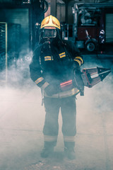 Portrait of a brave firefighter standing confident wearing full protective equipment, turnouts and helmet. Dark background with smoke and blue light.