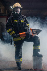 Fototapeta premium Portrait of a brave firefighter standing confident wearing full protective equipment, turnouts and helmet. Dark background with smoke and blue light.